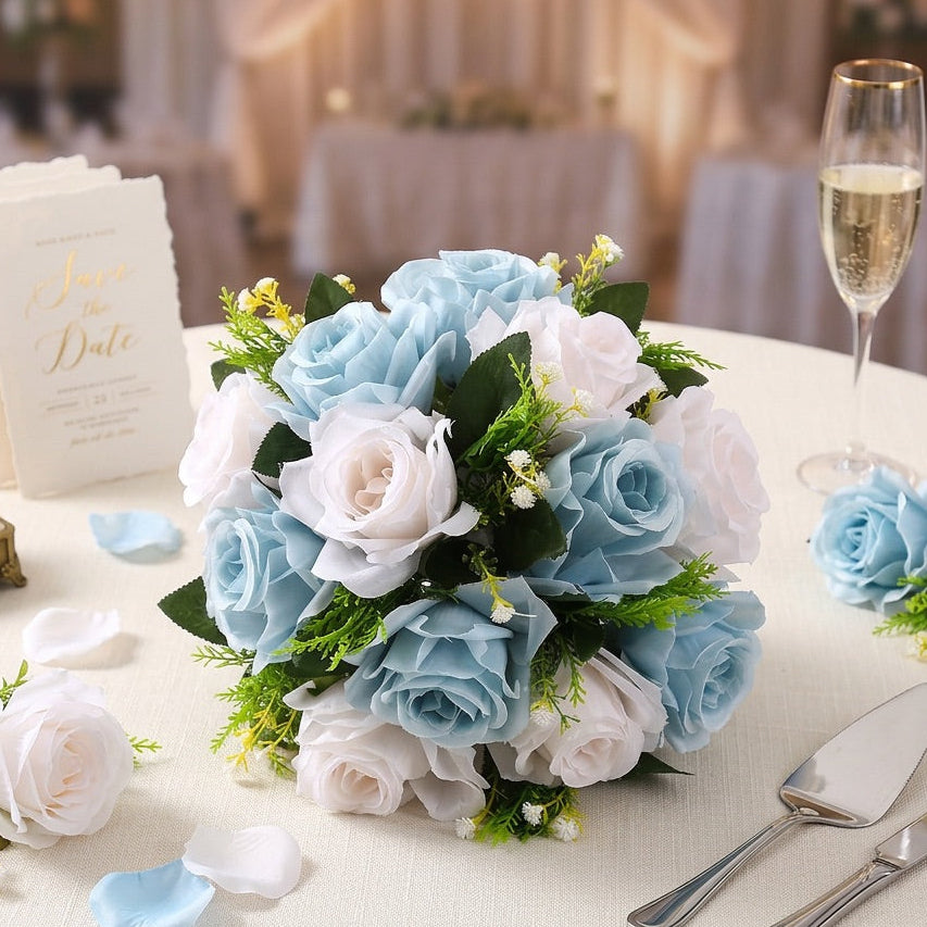 A round wedding table decorated with a central flower centrepiece of light blue and white roses, green leaves, and scattered petals, alongside champagne flutes, a save-the-date card, a small gold-toned box, and serving utensils.
