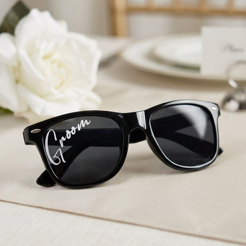 Black sunglasses with 'Groom' on the frame, placed on a white surface with white flowers in the background.
