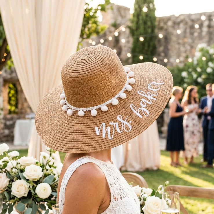 A woman at an outdoor wedding table wearing a wide-brimmed straw hat with white pom-pom trim and 'Mrs. Baker' written on the brim, paired with a white lace dress and holding a champagne flute.