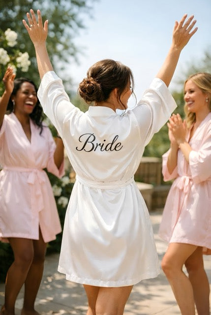 Four women wearing matching bridal party robes with ‘Bride’, ‘Maid of Honour’ and ‘Bridesmaid’ titles on the back, seen from behind.