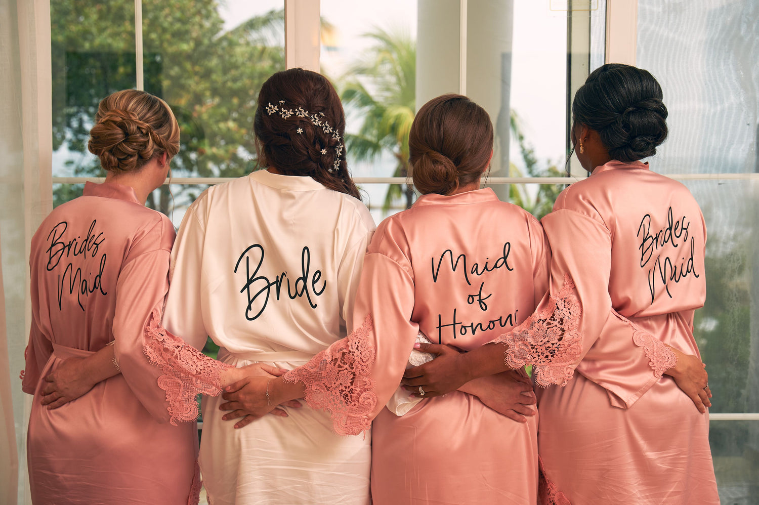 Four women wearing matching bridal party robes with ‘Bride’, ‘Maid of Honour’ and ‘Bridesmaid’ titles on the back, seen from behind.