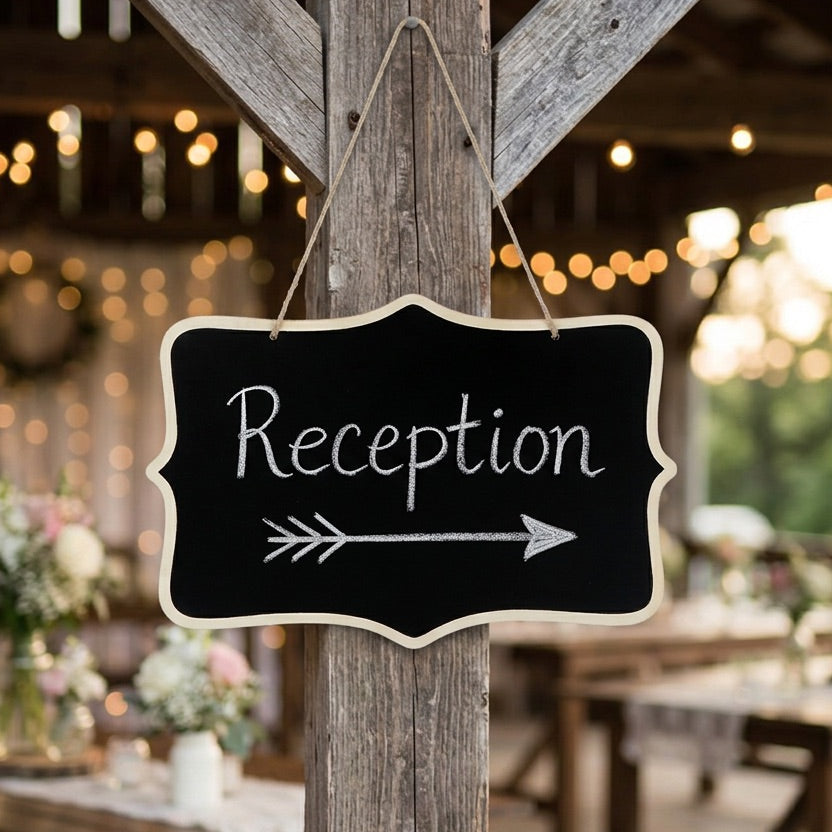 Black chalkboard sign with 'Reception' and an arrow hanging on a wooden post with a blurred background of a decorated indoor setting.