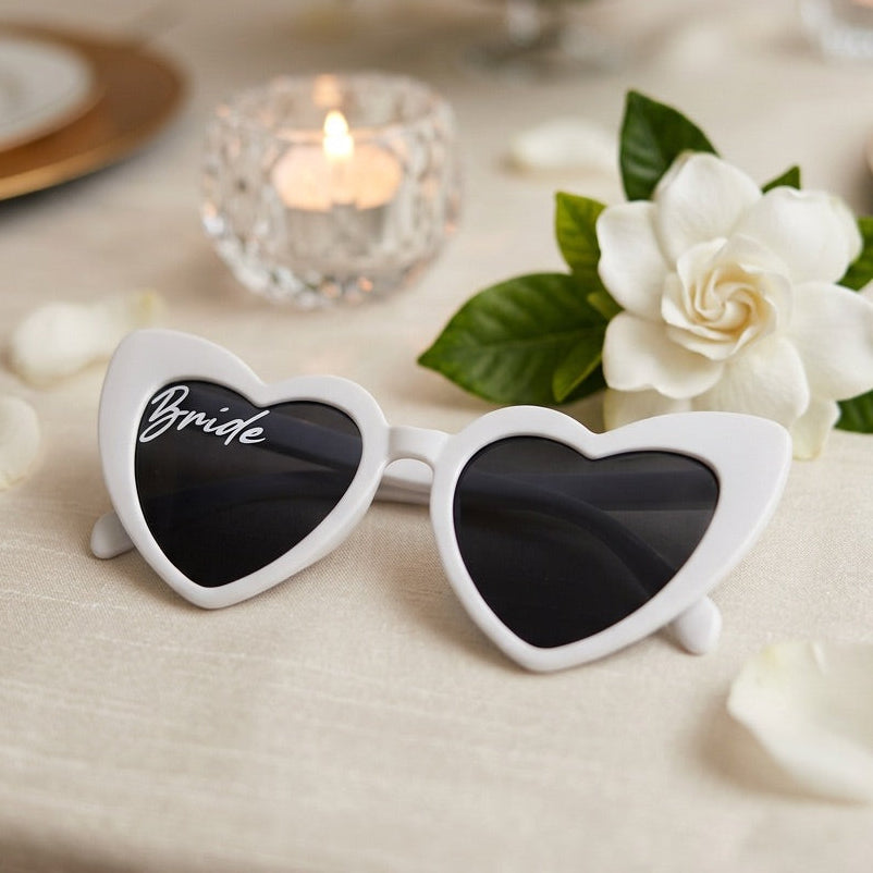 Heart-shaped sunglasses with 'Bride' on a table setting with flowers and candles.