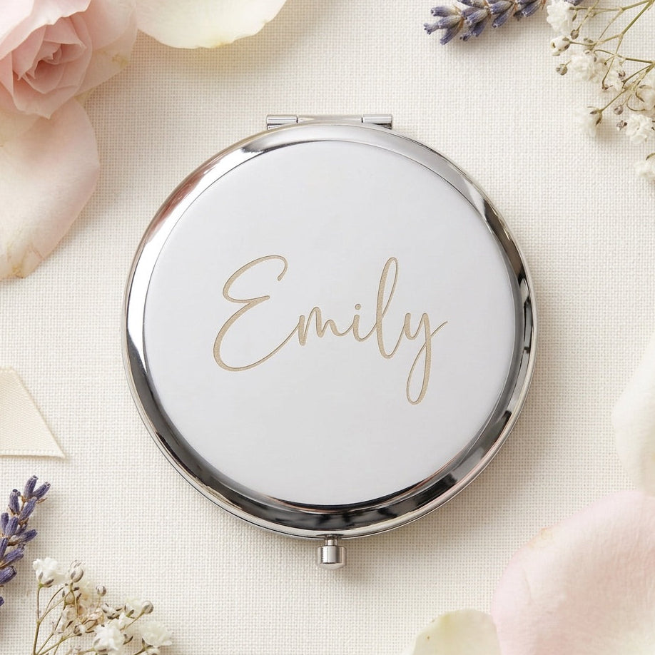 A round compact mirror with a polished silver frame and a white lid featuring the name 'Emily' in elegant gold script. The compact is styled in a flat-lay arrangement with soft pink and white roses and lavender sprigs on a textured cream fabric background.