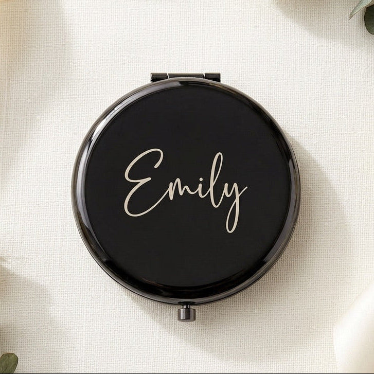 A round black compact mirror with the name 'Emily' written in elegant white script on the lid, styled in a flat lay with cream roses, eucalyptus stems, and cream ribbon on a textured off-white fabric background.