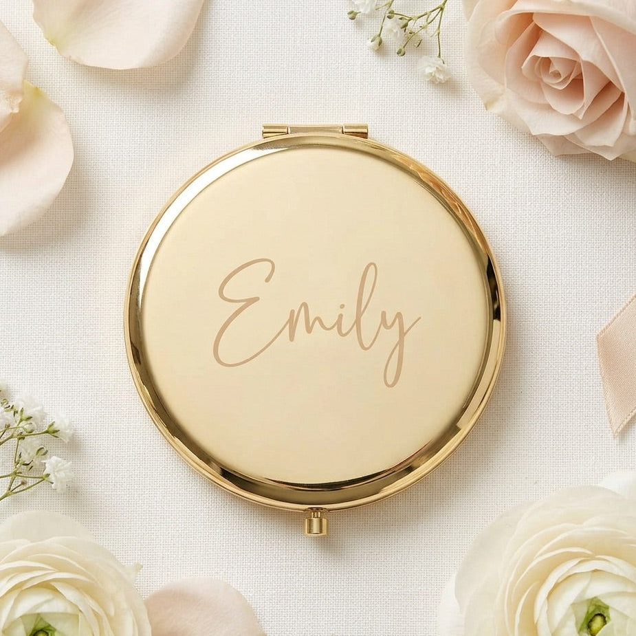 A round gold compact mirror with the name 'Emily' written in elegant gold script on the lid, styled in a flat lay with white and blush roses and baby's breath on a textured white fabric background.