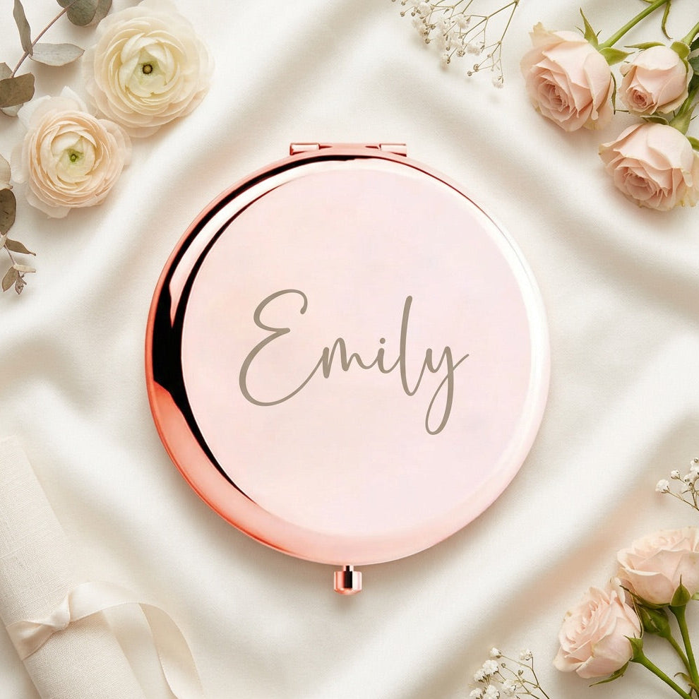 A round compact mirror with a rose gold metallic frame and a pale pink reflective surface, personalized with the name 'Emily' in gray script. The compact is styled in a flat-lay on draped white satin fabric, surrounded by soft pink roses and white baby's breath.
