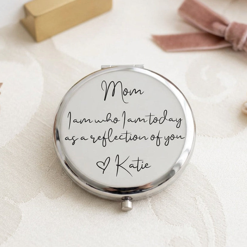 A round silver compact mirror with a white reflective surface, personalized with the text 'Mom I am who I am today as a reflection of you Katie' in black script. The compact is styled on a textured cream fabric with soft pink flowers, a gold box, and a pink ribbon in the background.