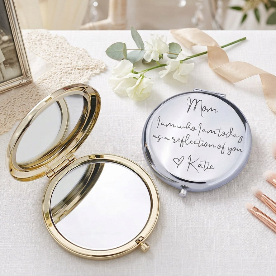 A round compact mirror with a gold-toned frame shown open beside another closed compact mirror engraved with the message 'Mom I am who I am today as a reflection of you Katie'. The scene is styled on a white surface with white flowers, a cream ribbon, makeup brushes, and a soft blurred interior background.