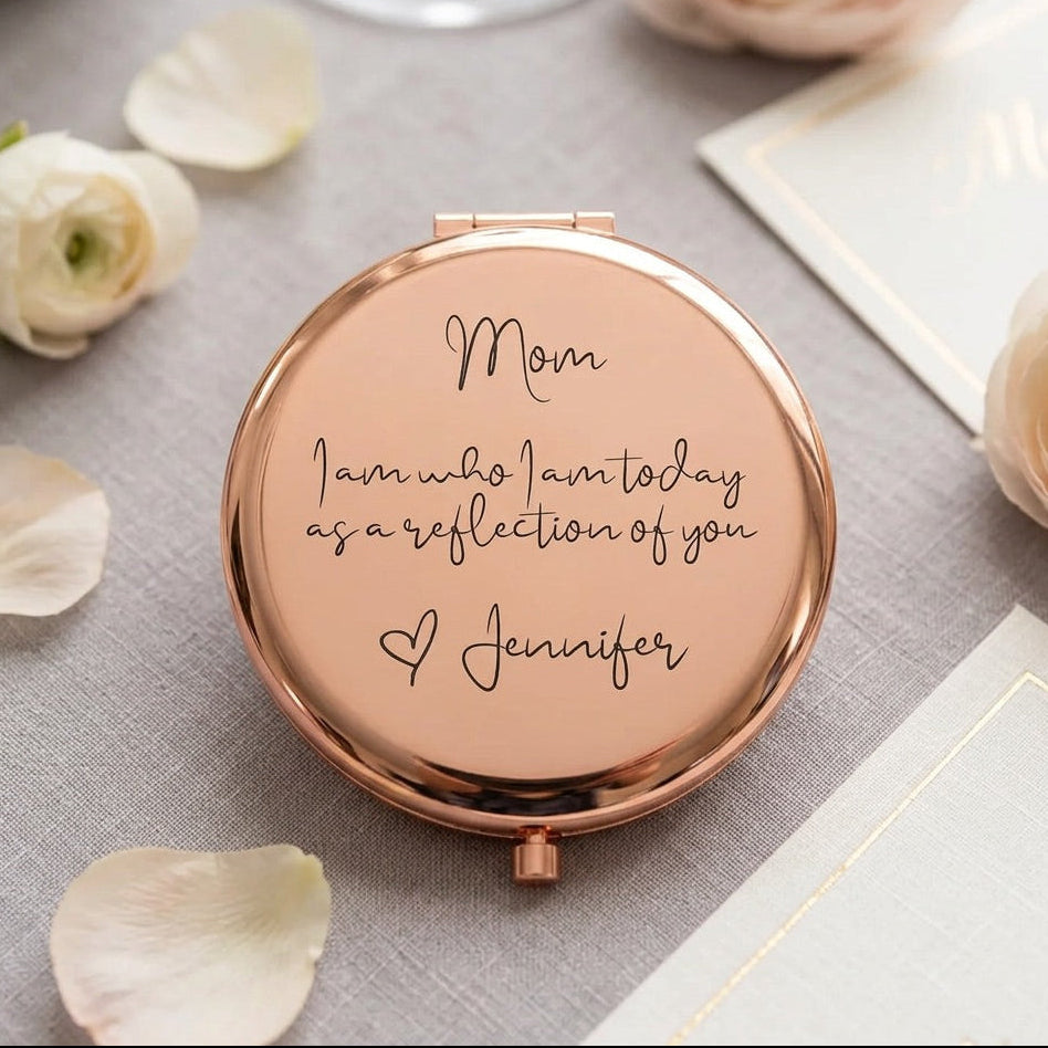A round rose gold compact mirror with a personalized message printed on the lid, displayed on a gray textured fabric with white rose petals and stationery.