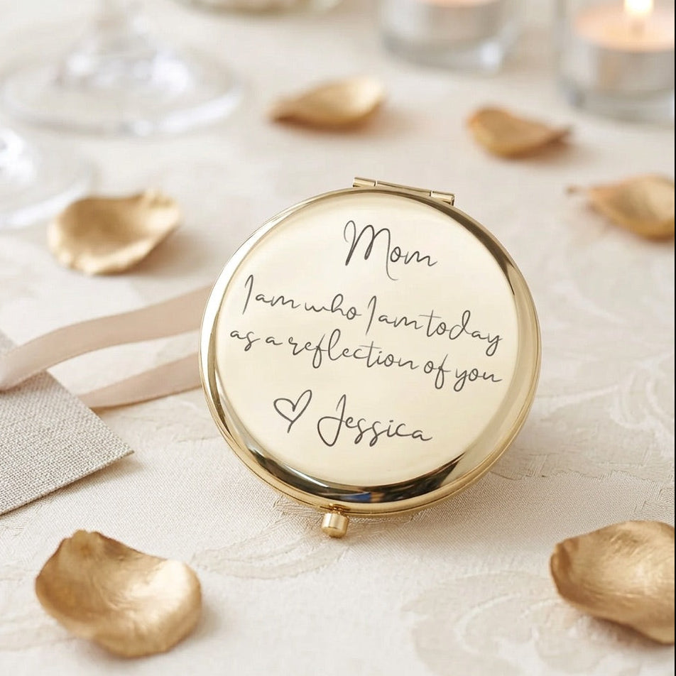 A round gold compact mirror with a cream-colored reflective surface, engraved with the message 'Mom I am who I am today as a reflection of you & Jessica' in black script. The compact is styled on a white textured fabric with scattered gold rose petals, a ribbon, and soft background bokeh.