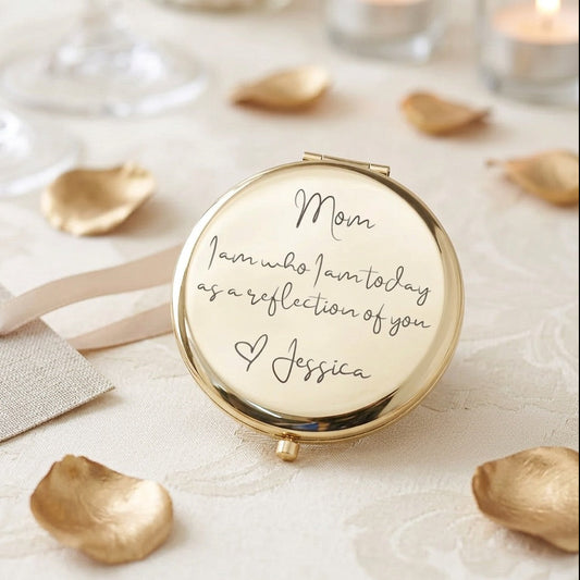 A round gold compact mirror with a cream-colored reflective surface, engraved with the message 'Mom I am who I am today as a reflection of you & Jessica' in black script. The compact is styled on a white textured fabric with scattered gold rose petals, a ribbon, and soft background bokeh.