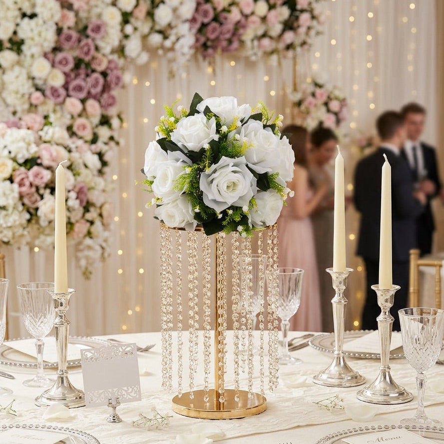 A wedding reception table setting featuring a centerpiece of white roses in a gold and crystal bead vase, surrounded by silver candlesticks, clear glassware, and white table linens, with a blurred background of floral arrangements and guests.