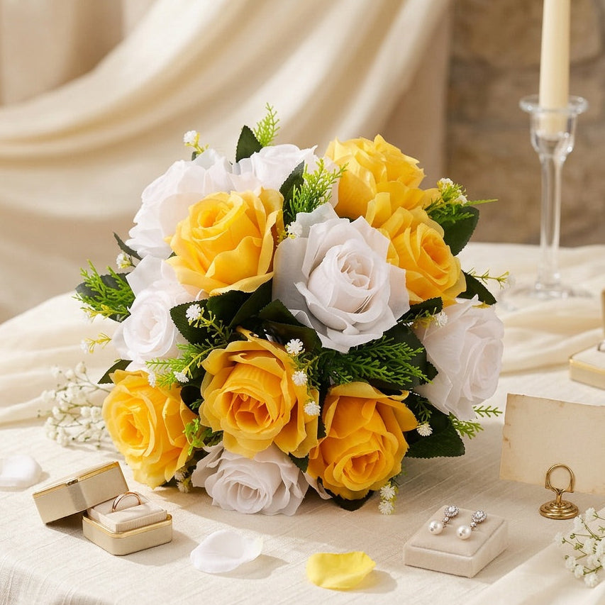 A centrepiece of yellow and white roses with green leaves arranged on a draped cream tablecloth, with small jewelry boxes and rings nearby and a candle holder in the background.