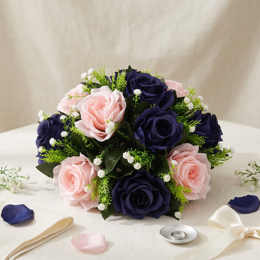 A flower centrepiece  of artificial roses in navy blue and light pink with green foliage, arranged on a white draped table with ribbon, petals, and a small ring holder.