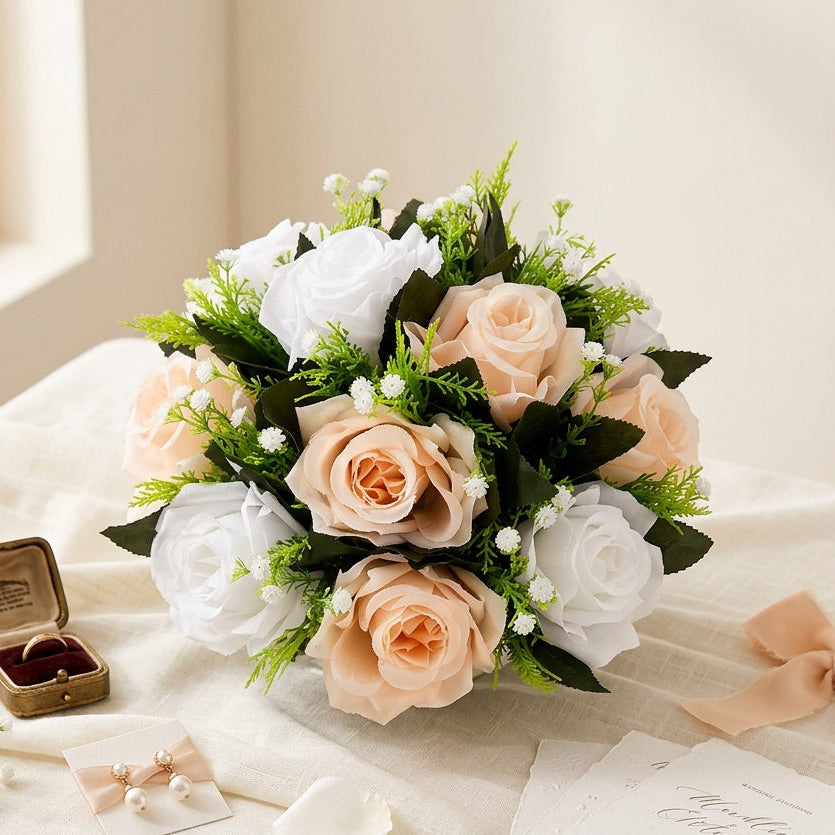 A centrepiece of faux roses in blush peach, white, and cream tones with green foliage, arranged on a cream tablecloth beside a ring box, invitation cards, petals, and a ribbon bow.