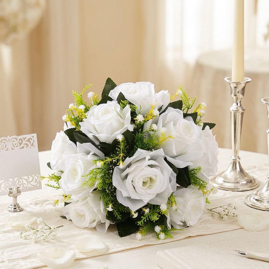 A flower centrepiece  of white artificial roses with green leaves and small yellow filler flowers, arranged on a cream table runner with silver candlesticks, a place card, and a pen in a softly lit indoor setting.