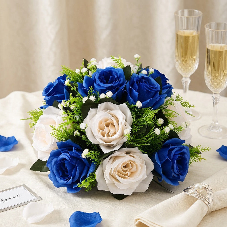 A centrepiece of blue and white faux roses with green foliage and small white blossoms, arranged on a cream tablecloth with champagne flutes, scattered petals, a folded napkin, and a place card.