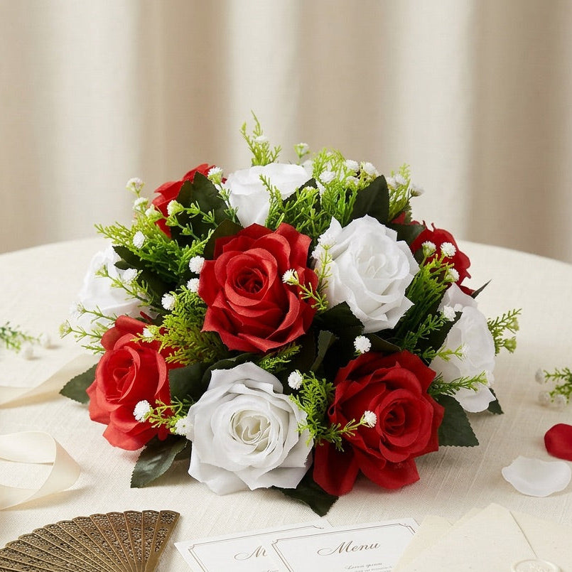 A round table set with a red and white rose flower centrepiece , cream stationery cards and envelopes, a gold fan, and scattered rose petals against a soft beige curtain background.