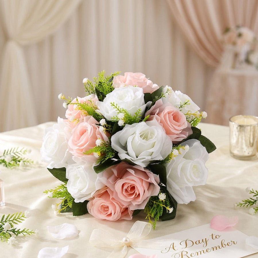 A round artificial flower centerpiece arrangement of blush pink and white roses with green foliage, displayed on a cream draped table with candles, petals, and a 'A Day to Remember' card.