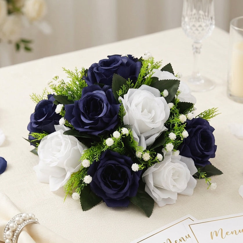 A flower centrepiece  of artificial navy blue and white roses with green foliage and small white filler flowers, arranged on a cream tablecloth with wedding-themed props including a napkin, menu cards, petals, and a candle.