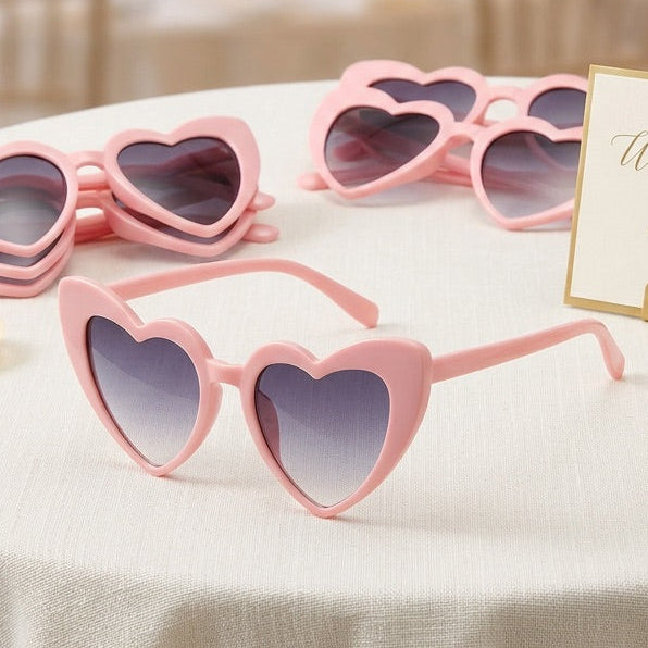 Pink heart-shaped sunglasses on a table with a decorative sign and flowers in the background.