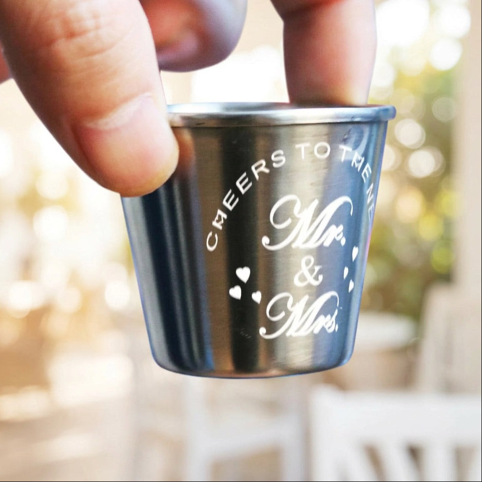Metal shot glass with 'Cheers to the Mr. & Mrs.' text held by a hand against a blurred outdoor background.
