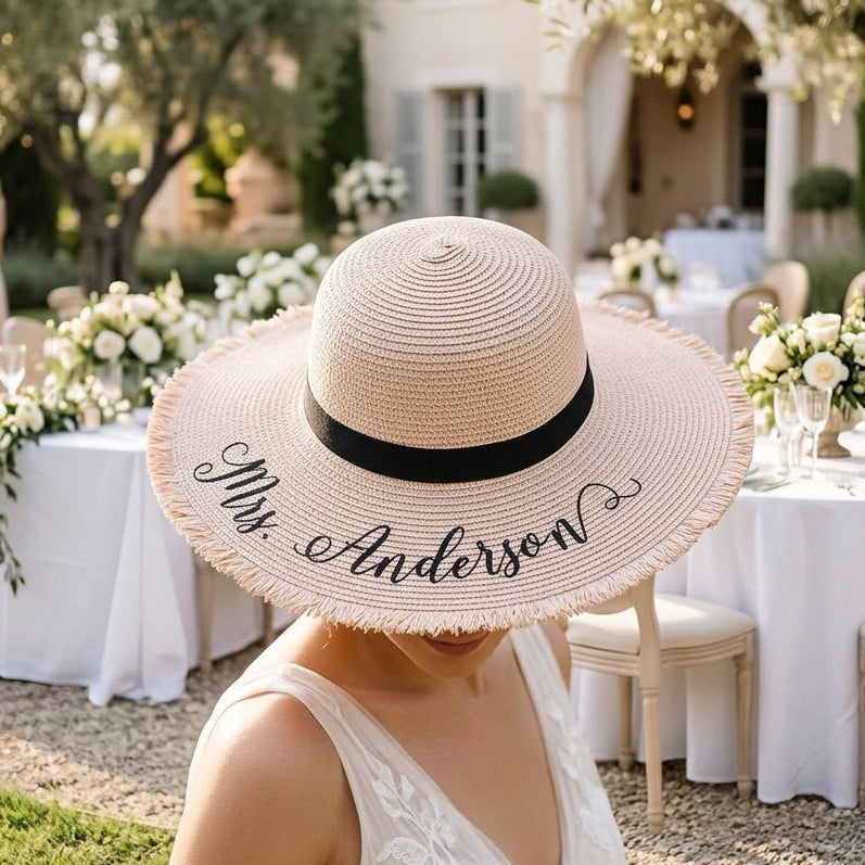 A woman outdoors at a wedding reception wearing a large woven straw sun hat with a black band and the words 'My Anderson' embroidered on the front. White tables, greenery, and guests are blurred in the background.