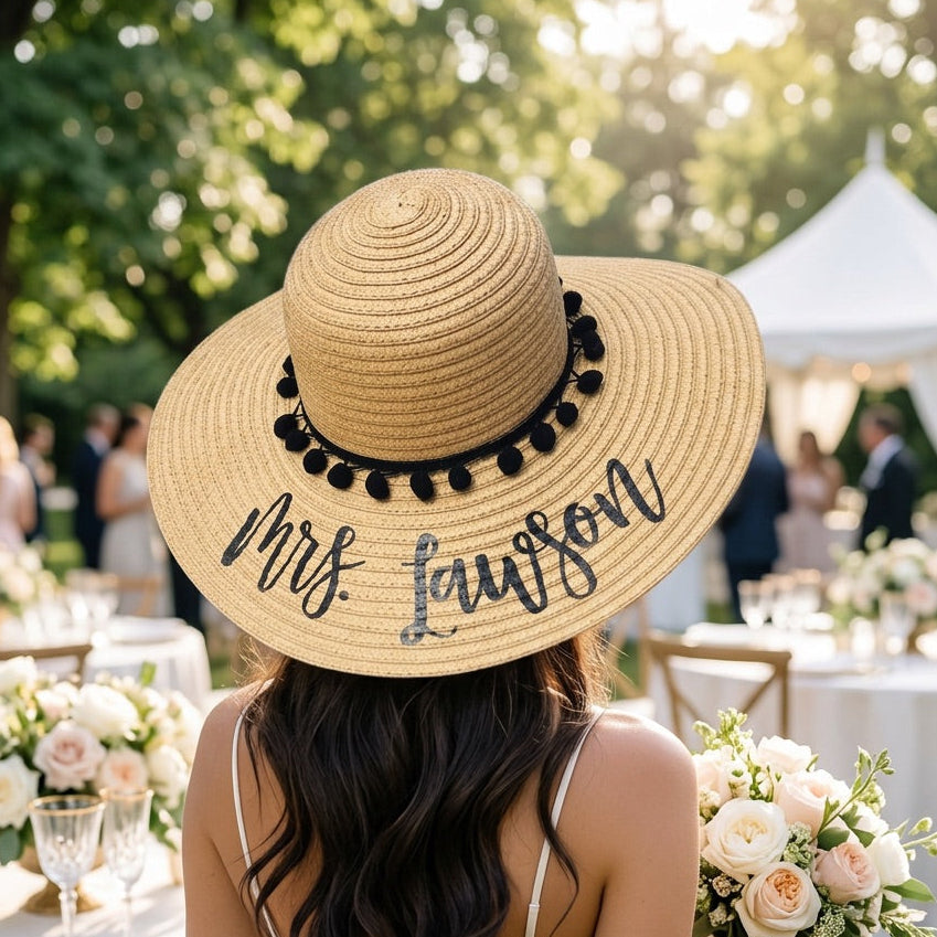 A woman seen from behind at an outdoor wedding, wearing a large natural straw sun hat with black pom-pom trim and the name 'Mrs. Lawson' written in black script across the brim.
