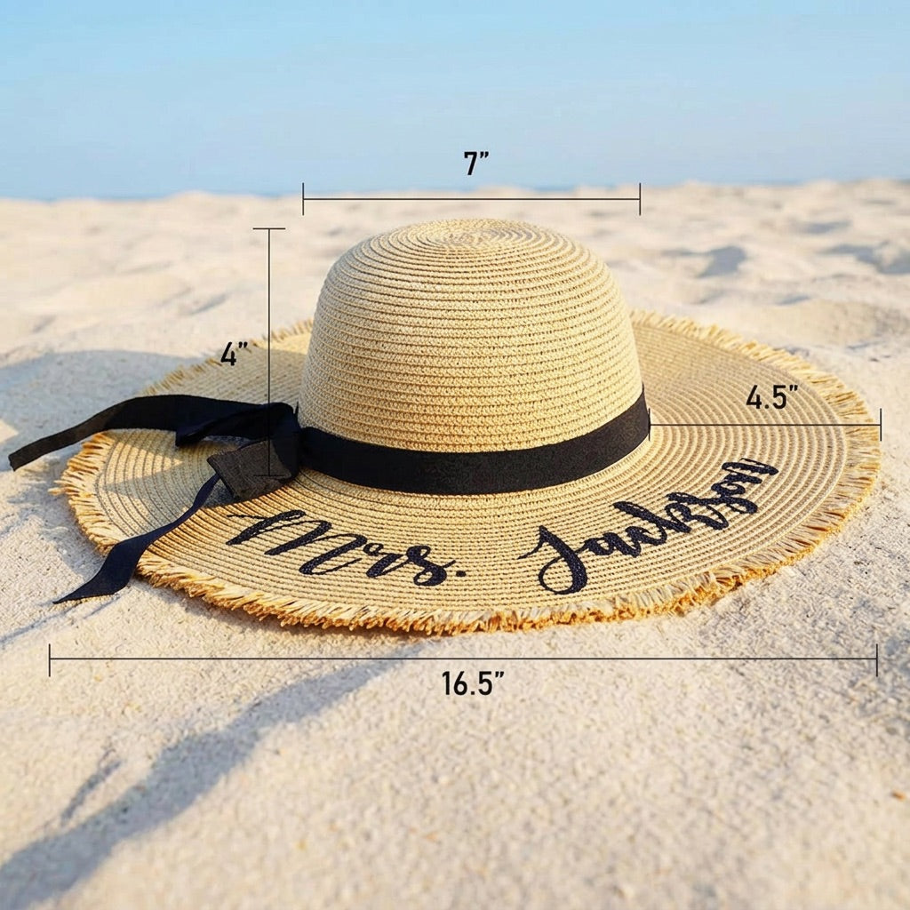 A straw sun hat with a black ribbon and black script lettering on the brim, photographed on sand with overlaid dimension lines and measurements.