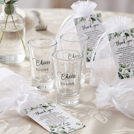 Three clear glass cups printed with 'Cheers TO LOVE' arranged on a beige patterned tablecloth, alongside white drawstring gift bags and printed thank-you cards with greenery graphics. A glass vase with eucalyptus stems sits in the background, and warm blurred lights appear out of focus behind the setup.