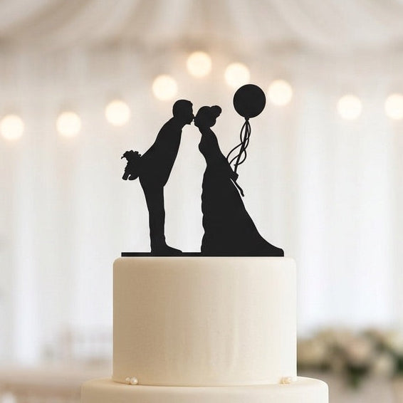 Three-tiered white wedding cake with black silhouette topper in a decorated room.