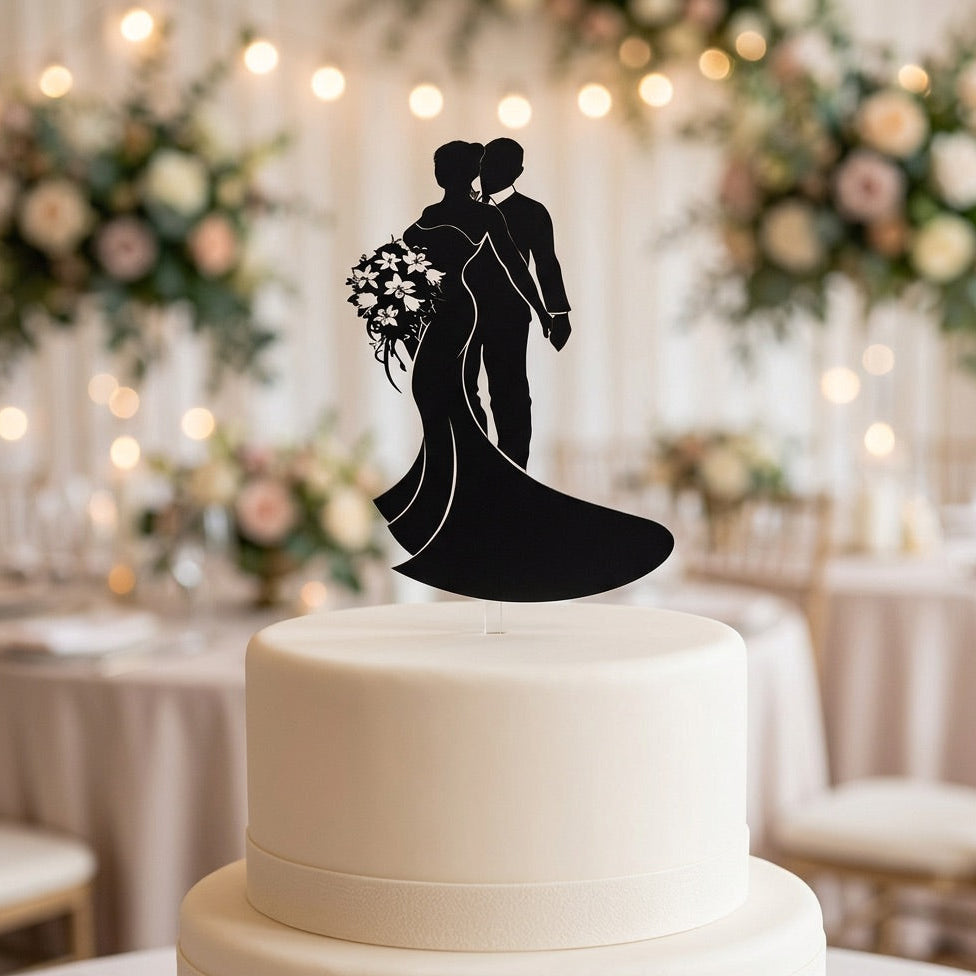 Silhouette cake topper of a bride and groom on a white cake with a blurred wedding reception background.