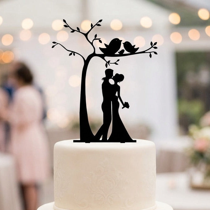 White wedding cake with a black silhouette topper of a couple under a tree, with blurred guests and lights in the background.