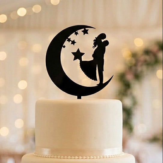 White wedding cake with a decorative topper featuring a couple on a moon and star design, set against a blurred wedding reception background.